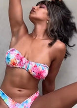 Load image into Gallery viewer, A strapless bikini top with a ruched design in multicolor floral pattern, being modeled in a seated pose by a woman.