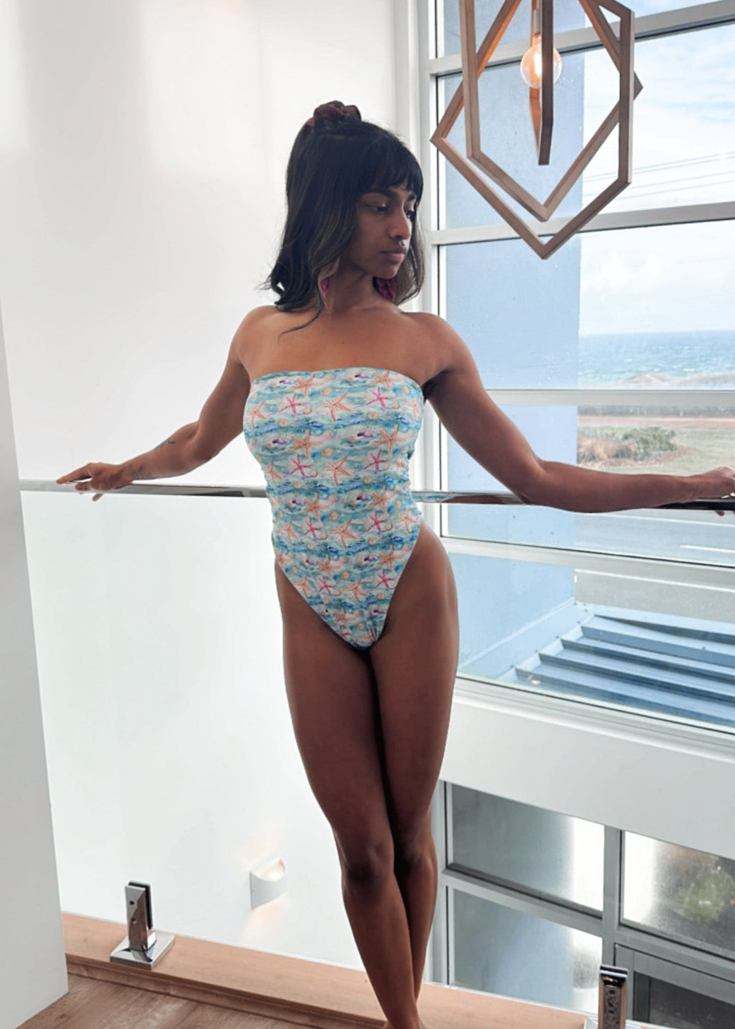 Woman in a floral swimsuit standing by a large window with a cityscape view.